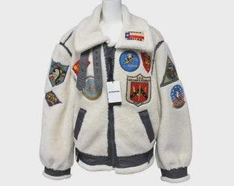 Könnte beinhalten: Cremefarbene Jacke im Shearling-Stil mit mehreren gestickten Aufnähern. Verfügt über einen breiten Kragen, dunkelgraue Zierleisten und einen Reißverschluss. Aufnäher mit Flaggen, Emblemen und Text. Ein Etikett mit dem Wort "ATTENTION" hängt am Reißverschluss.