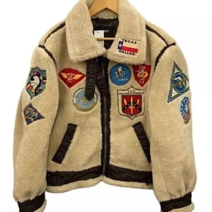May include: A tan fleece jacket with a shearling collar and multiple embroidered patches. The jacket has a black zipper, black trim, and a "Texas Dallas" patch. The jacket is hanging on a wooden hanger.