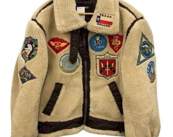 Könnte beinhalten: Beige Fleecejacke mit Schafsfellkragen und mehreren gestickten Aufnähern. Die Jacke hat einen schwarzen Reißverschluss, schwarze Paspeln und einen "Texas Dallas" Aufnäher. Sie hängt an einem Holzkleiderbügel.