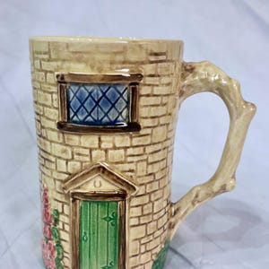 May include: A novelty ceramic mug designed as a stone tower, with a green door, blue window, and a handle shaped like a branch. The beige mug has brown detailing and pink floral accents, evoking a charming, rustic style.