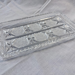 May include: A rectangular clear glass tray featuring a pressed geometric pattern with starburst motifs and a scalloped edge. The tray has a divided interior section and a faceted rim, reflecting light through its intricate, textured surface design.