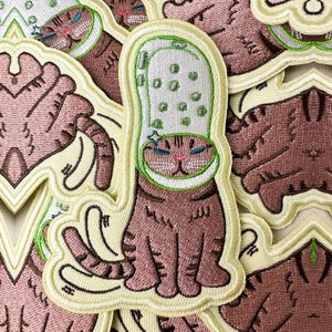 Puede incluir: Parche bordado con un gato atigrado marrón que lleva un zapato blanco y verde. El gato tiene ojos azules y nariz rosa. El parche tiene un borde amarillo, ideal para ropa o accesorios.