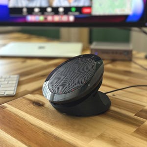 May include: A black and silver conference speakerphone on a wooden desk. The speakerphone has a circular speaker grill and is connected to a power source. A computer monitor displaying a video call is in the background.