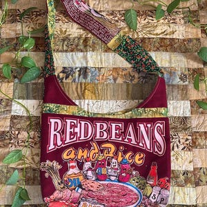 May include: A burgundy shoulder bag with a colorful graphic of red beans and rice. The bag features a patterned strap with floral and text details. The text "REDBEANS and Rice" is prominently displayed on the front.