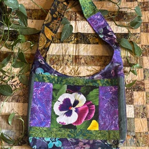 May include: A handmade, quilted shoulder bag with a large pansy flower design. The bag features a patchwork design with purple, green, and black fabrics. The strap has a floral pattern. The bag is approximately 15 inches wide and 12 inches tall.