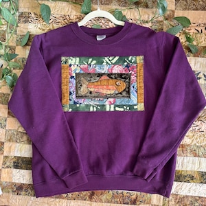 May include: A deep purple sweatshirt with a rectangular patch featuring a fish design. The fish is orange and brown, set against a backdrop of floral and patterned fabrics. The sweatshirt is hanging on a white hanger.
