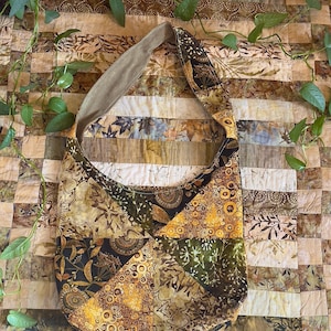 May include: A patchwork shoulder bag with a beige strap and a patterned body. The bag's design incorporates various earth-toned fabrics with floral and geometric motifs. The background is a quilt with similar colors.