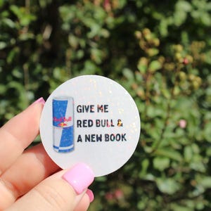 May include: A round sticker with the text "GIVE ME RED BULL & A NEW BOOK" and a Red Bull can illustration. The sticker has a glittery, white background and is held in front of a blurred green background.