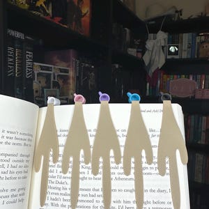 May include: A row of beige, hand-shaped bookmarks with colorful tops are inserted into the pages of an open book. The bookmarks have a unique design, with each one featuring a different color on top. The book is surrounded by a bookshelf.