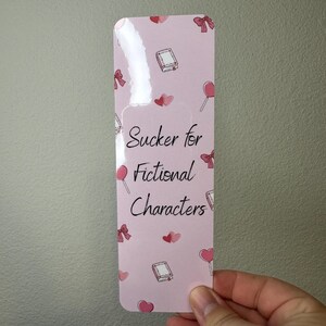 May include: A pink bookmark with the text "Sucker for Fictional Characters" in black script. The bookmark is decorated with small illustrations of books, bows, hearts, and lollipops. The bookmark is held by a hand.