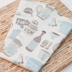 Puede incluir: Dos toallas de cocina blancas con un patr&oacute;n azul y marr&oacute;n de utensilios de cocina, incluyendo un batidor, cucharas medidoras, una jarra y una taza de t&eacute;. Las toallas est&aacute;n dobladas y colocadas sobre una superficie de madera.