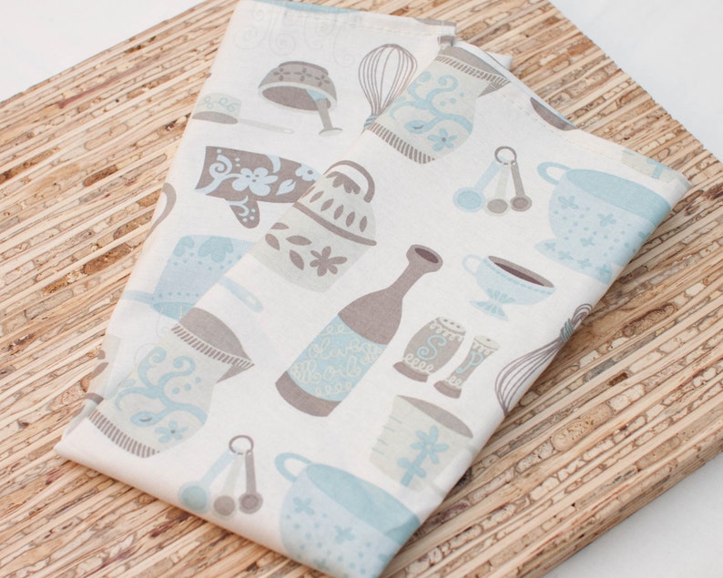 Puede incluir: Dos toallas de cocina blancas con un patr&oacute;n azul y marr&oacute;n de utensilios de cocina, incluyendo un batidor, cucharas medidoras, una jarra y una taza de t&eacute;. Las toallas est&aacute;n dobladas y colocadas sobre una superficie de madera.