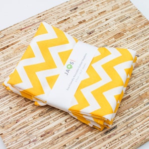 Puede incluir: Un montón de servilletas de algodón con estampado de chevrón amarillo y blanco, sujetas por una banda blanca con texto. Las servilletas miden 41,9 cm cuadrados. Las servilletas están sobre una superficie de madera marrón claro.