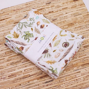 Puede incluir: Un paquete de cuatro servilletas de algodón blanco con un estampado de temática natural que presenta bellotas, hojas, plumas y una rana. Las servilletas miden 42 cm cuadrados y están envueltas con una banda blanca.