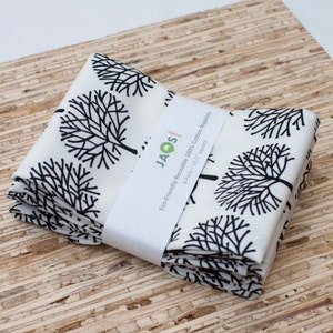 Puede incluir: Un paquete de cuatro servilletas de algodón blanco con un diseño de árbol negro. Las servilletas están dobladas y empaquetadas con una etiqueta que dice "Servilletas de algodón 100% reutilizables ecológicas 4 unidades - 16,5" x 16,5" - Jaos"