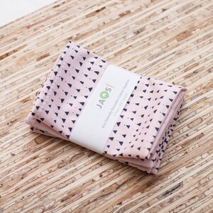 May include: A stack of four pink reusable cotton napkins with a navy blue triangle pattern. The napkins are wrapped with a white label that reads "JAQS" and "Eco-Friendly Reusable Cotton Napkins". Each napkin is 12 x 12 inches.
