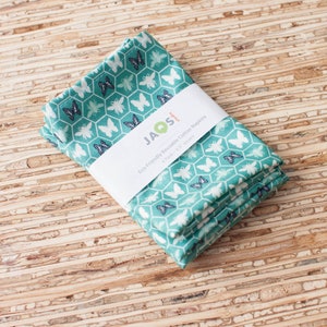 以下が含まれることがあります： 白い蝶と蜂の模様がついた、ターコイズブルーの綿製ナプキンが4枚重ねられています。ナプキンは、「JAOS 環境に優しいリサイクルコットンナプキン 4パック - 17 x 17インチ」と書かれた白いラベルで包装されています。