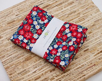 Op de afbeelding: Een vierpak herbruikbare katoenen servetten met een donkerblauwe achtergrond en een dicht patroon van rode, witte en lichtblauwe bloemen met groene bladeren. Een witte papieren band bevat de tekst JAO studio 4 Pack - 41,9 cm vierkant.