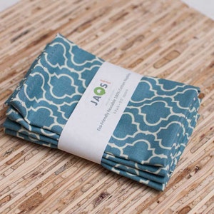 May include: A stack of four teal and white patterned cotton napkins, each measuring 9.5 inches square, wrapped with a white band that reads "JAQS studio" and "Eco-Friendly Reusable 100% Cotton Napkins". The napkins are folded and stacked neatly.