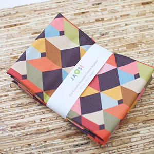 May include: A stack of four folded, colorful cotton napkins with a geometric cube pattern in shades of orange, pink, green, and brown. The napkins are wrapped with a white band that reads "JAOS! Eco-Friendly Reusable Cotton Napkins."