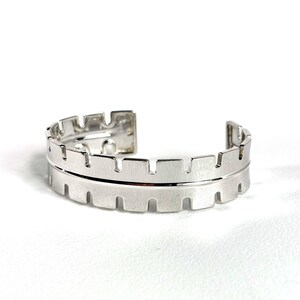 May include: A silver-toned cuff bracelet featuring a notched, gear-like edge design along both the top and bottom borders. The metal surface has a brushed finish, and the piece is open at the back, forming a circular shape with a central horizontal line.