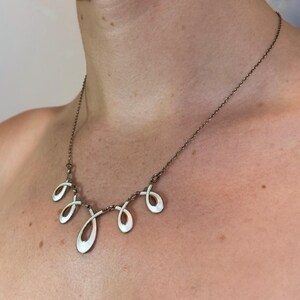 Peut inclure: Un collier argenté composé d'une chaîne fine et d'un pendentif central formé de cinq boucles en forme de goutte. Chaque boucle est remplie d'émail blanc et présente de fines lignes gravées rayonnantes sur toute sa surface.