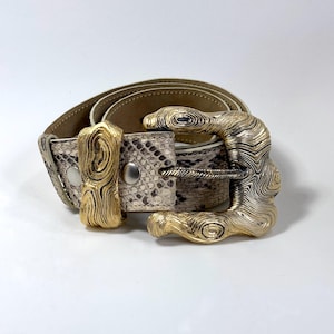 Puede incluir: Un cinturón con estampado de piel de serpiente y hebilla dorada. El cinturón presenta una hebilla dorada texturizada y una trabilla a juego. El cinturón está hecho de un material con estampado de piel de serpiente con una paleta de colores neutros.