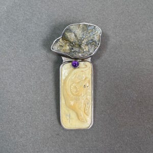 May include: A silver pendant with an ivory-coloured rectangular base, featuring a carved ram design. A purple gemstone is set above the ram, and a rough-cut stone tops the pendant. A unique piece of jewellery.