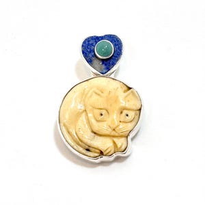 May include: A cream-coloured pendant with a carved cat design, set in a silver frame. Above the cat is a blue heart-shaped stone with a small, round, green stone in the centre. The pendant is likely a piece of jewellery.