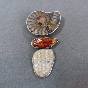 Puede incluir: Tres colgantes de fósiles con bordes de metal plateado. El colgante superior es un amonita marrón y beige. El colgante del medio es una piedra pulida de color naranja-marrón. El colgante inferior es un fósil color crema con un patrón ondulado.