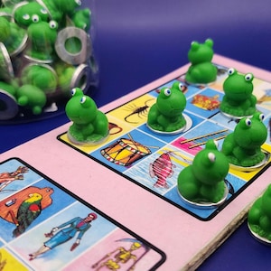 May include: A board game with green frog game pieces. The game board is pink with colorful illustrations. A jar filled with additional frog pieces sits in the background. The frogs are on small silver bases.