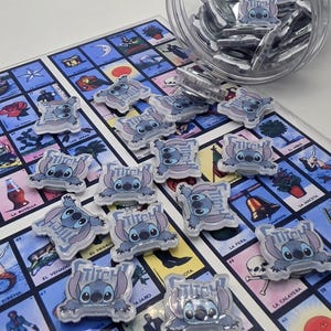 May include: Clear acrylic Stitch character charms scattered on a colorful game board. The charms feature a light blue Stitch design with the word "Stitch" above. A clear container holds additional charms. The game board has various illustrations and text.