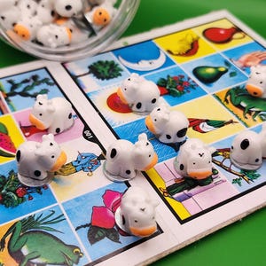 May include: A close-up of a traditional Mexican Loteria game. The game board features colorful illustrations, with small white and orange cow figurines placed on the board. A clear jar filled with similar figurines is in the background.