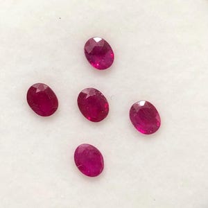 May include: Five loose, oval-cut rubies with a deep red colour and faceted surfaces. The gemstones are arranged in a cross-like pattern on a white foam insert inside a clear plastic display case, showcasing their clarity and vibrant red hue.