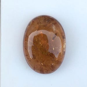 May include: An oval-shaped, polished gemstone with a translucent, warm amber-brown color. The stone features a smooth, rounded cabochon surface that reflects light, revealing internal natural inclusions and variations in its golden-brown hue.