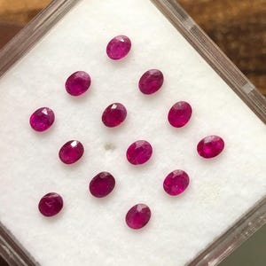 May include: A collection of twelve small, oval-shaped, faceted rubies with a deep pinkish-red colour. The gemstones are arranged in a grid pattern on a white foam insert inside a clear, square plastic display case with a hinged lid.