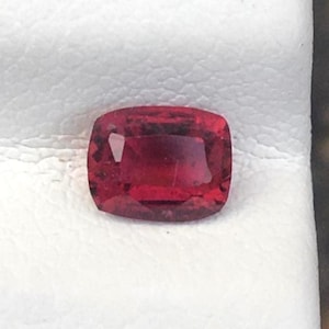 Puede incluir: Una piedra preciosa de corte cojín con un color rojo profundo y saturado y una superficie facetada que refleja la luz. La piedra tiene una forma rectangular con esquinas redondeadas y muestra claridad interna con sutiles inclusiones naturales.