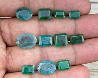 May include: An assortment of emerald and aquamarine gemstones in various shapes, including oval and rectangular cuts. The gems display shades of green and blue, with visible facets. The arrangement showcases the different sizes and cuts of the gemstones.