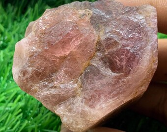 May include: A rough-cut, pink gemstone with a crystalline structure. The stone has a mix of light and dark pink hues, with some areas appearing translucent. The surface is uneven and textured, with visible ridges and imperfections.