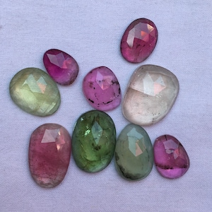 May include: An assortment of faceted gemstones in various colors, including pink, green, and clear. The stones are oval-shaped and arranged on a white fabric surface. The gems have a polished, reflective surface.