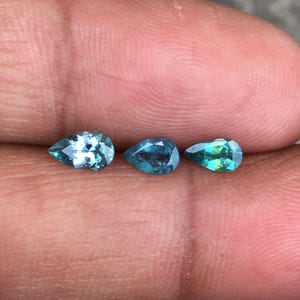 以下が含まれることがあります： 青とティール色の3つのペアシェイプの宝石。宝石はニュートラルな背景に表示され、そのファセットと内部の色のバリエーションが示されています。宝石はおそらくジュエリー作りのためです。