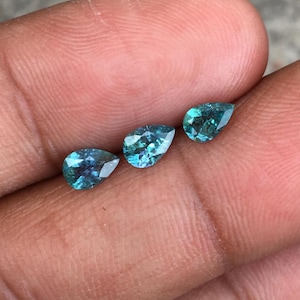 以下が含まれることがあります： 3つのティール色のペアシェイプの宝石。宝石はファセット加工された表面を持ち、光を反射してキラキラとした効果を生み出しています。石はニュートラルな背景に表示されています。