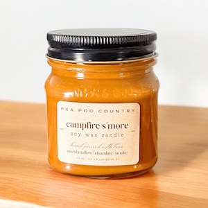 May include: A jar candle with a black lid and a label that reads "Campfire S'more." The candle is filled with a light brown wax and is 213 g. The label also says "Pea Pod Country" and lists the scents as marshmallow, chocolate, and smoke.