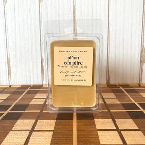 May include: A rectangular, light brown candle wax melt in a clear plastic clamshell package. The label reads "PEA POD COUNTRY piñon campfire - coconut soy wax candle - hand poured with love pine vanilla smoke". The candle weighs 2.5 oz.