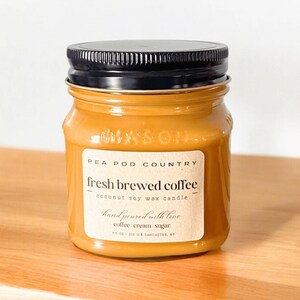 Peut inclure: Une bougie en pot avec un couvercle noir et une étiquette indiquant "fresh brewed coffee". La bougie est remplie d'une cire brun clair et porte l'étiquette "Pea Pod Country". L'étiquette indique également qu'il s'agit d'une bougie en cire de soja à la noix de coco, coulée à la main avec amour.