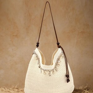 Könnte beinhalten: Cremefarbene gewebte Handtasche mit braunem Lederriemen. Die Tasche hat eine dekorative Verzierung mit baumelnden silberfarbenen Charms. Der Kordelzugverschluss ist braun, vor neutralem Hintergrund.