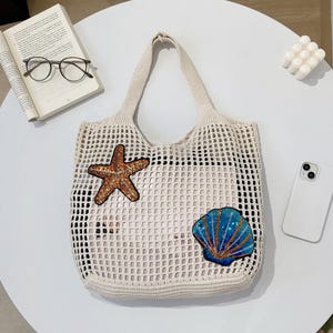 Könnte beinhalten: Cremefarbene Netztasche mit einem Seestern- und Muschelschmuck. Der Seestern ist braun und goldfarben, und die Muschel ist blau und goldfarben. Die Tasche hat eine rechteckige Form und zwei Henkel.