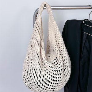 Könnte beinhalten: Eine cremefarbene, offen gewebte Tragetasche mit runder Form und einem dicken, gestrickten Griff. Die Tasche hängt an einer Metallstange, im Hintergrund ist eine schwarze Jacke zu sehen. Das Design der Tasche weist ein großes, offenes Netzmuster auf.