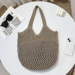 Könnte beinhalten: Beigefarbene, offen gewebte Tragetasche mit rundem Boden und zwei Henkeln. Die Tasche liegt auf einem weißen Tisch neben einer Zeitung mit dem Text "BELIEVE YOU CAN AND YOU ARE HALFWAY THERE" und einer Sonnenbrille.