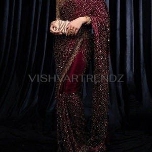 May include: A deep burgundy saree adorned with detailed gold embroidery, complemented by a gold necklace and bracelet. The saree displays a geometric design and is accompanied by a small, patterned clutch. The text "VISHVARTRENDZ" is visible.